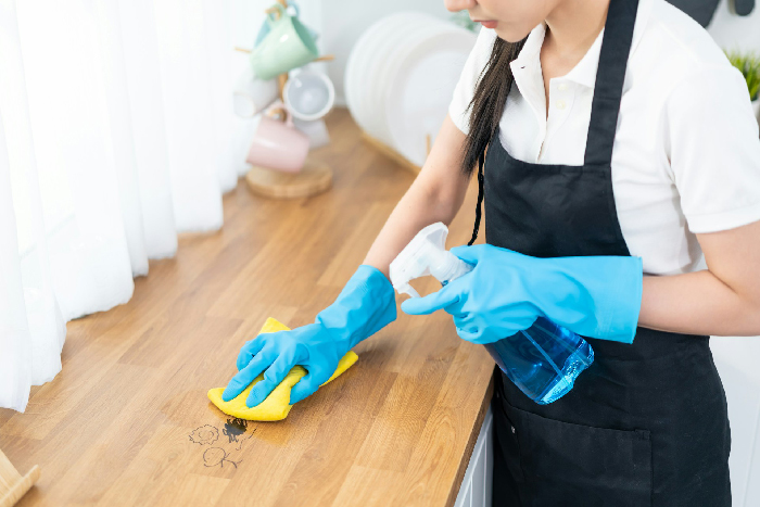 Woman wearing blue gloves and black apron cleaning a wooden countertop with a spray bottle and yellow cloth.