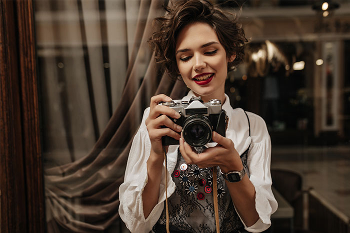 Young woman holding a camera, smiling indoors, illustrating man catching a friend stealing his boyfriend&rsquo;s camera scenario.