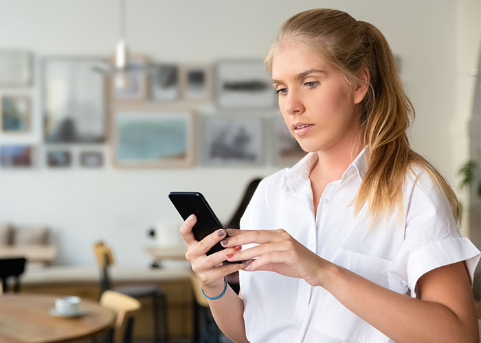 Young woman in white shirt looking at phone, appearing thoughtful about friends husband dating the same guy situation.