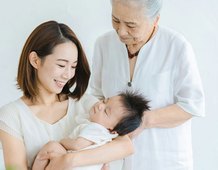 Young woman smiling while holding a baby and an elderly woman watching, illustrating neighbor mom spying and fence holes.