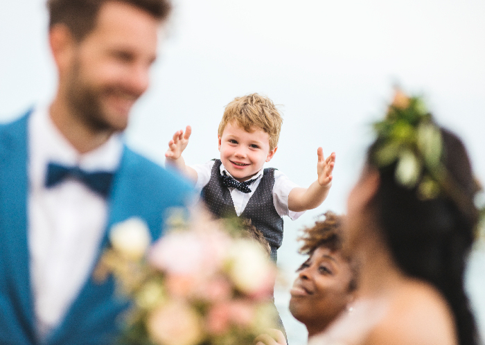 Young boy in a bow tie reaching out as bride and groom smile during an outdoor wedding ceremony. Young boy in a bow tie reaching out as bride and groom smile during an outdoor wedding ceremony.