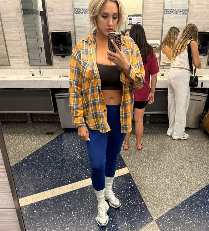 Model taking mirror selfie in leggings and open flannel, highlighting big breasts discrimination by flight attendant controversy.