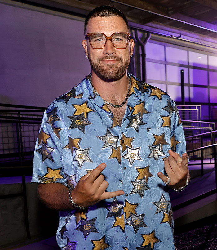 Travis Kelce wearing star-patterned shirt and clear glasses, posing casually as fans react to Taylor Swift video answer.