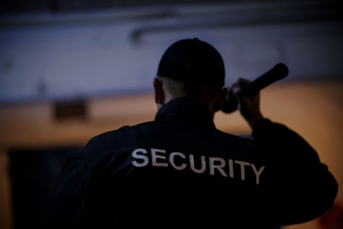 Security guard in dark uniform holding flashlight, inspecting a dimly lit area during a nighttime shift on the job.
