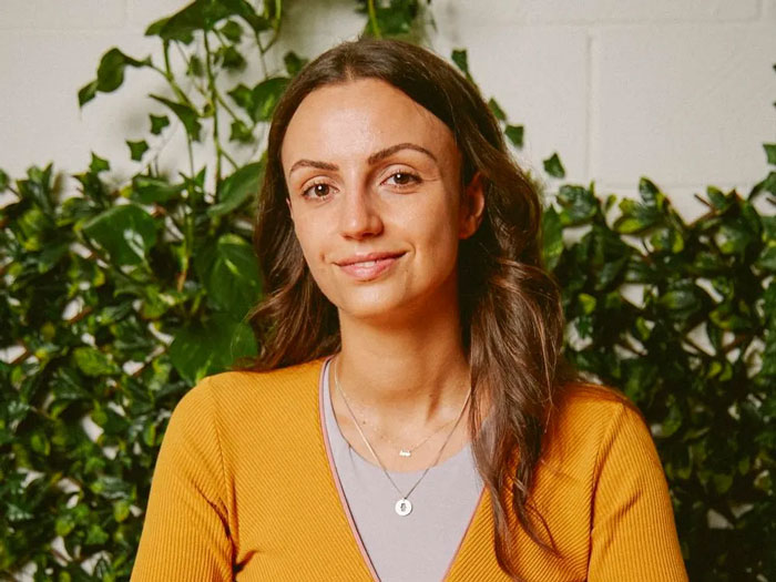 Woman with long brown hair wearing a mustard sweater, smiling calmly in front of green leafy plants indoors.