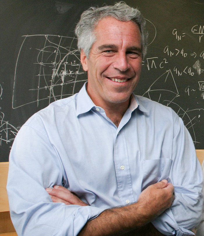 Jeffrey Epstein smiling in a light blue shirt standing in front of a blackboard with scientific equations and diagrams.