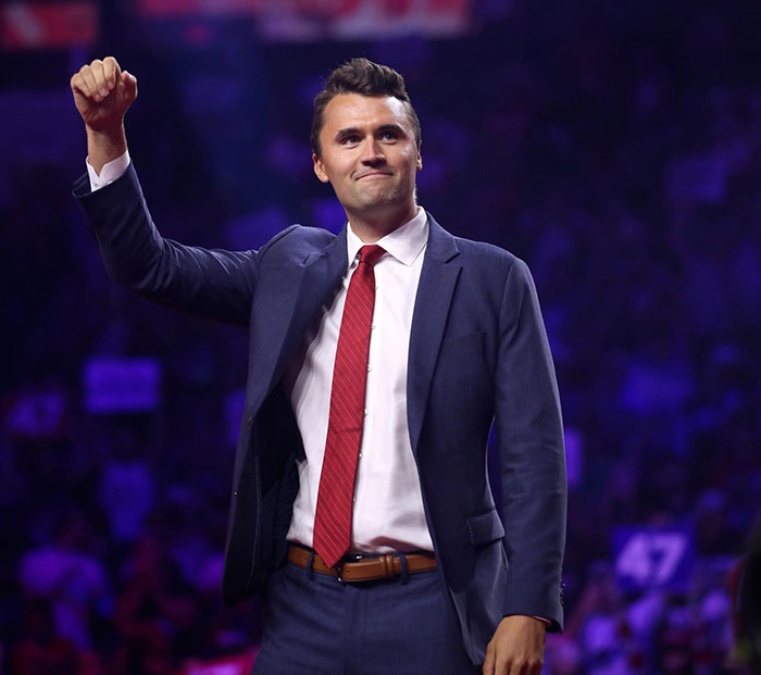 Charlie Kirk in a dark suit and red tie raising his fist at a crowded event, highlighting Charlie Kirk's wife haunting post.