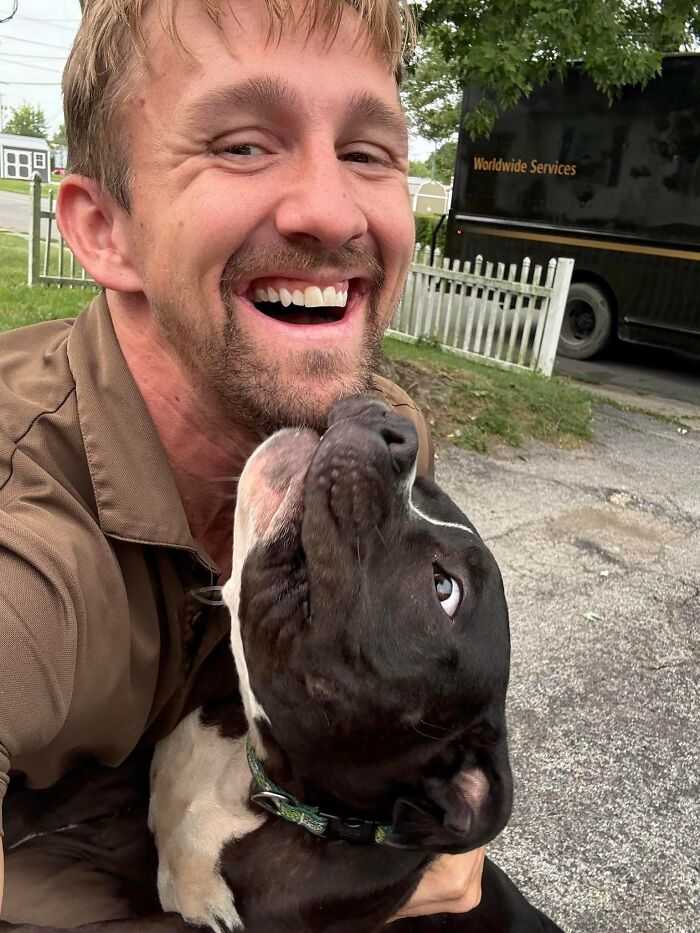 UPS driver smiling and happily hugging an adorable black and white dog outside near a delivery truck and white fence.