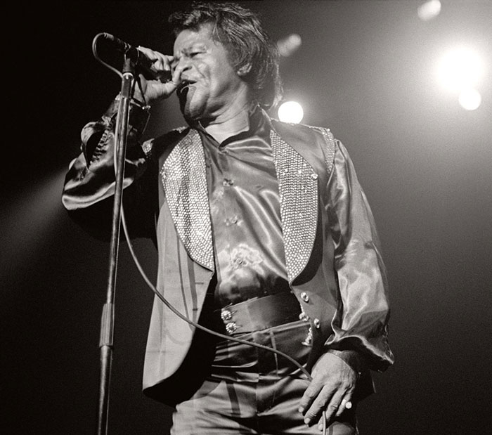 Black and white image of a male singer performing on stage at a concert with bright lights in the background.