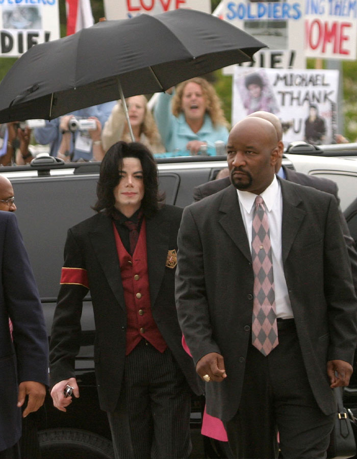 Michael Jackson walking with security, surrounded by fans and signs, highlighting intimate life stories by Priscilla Presley.