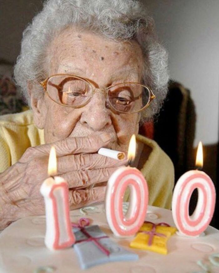 Elderly woman celebrating 100th birthday while lighting cigarette near birthday cake with candles, street photo capturing old age.