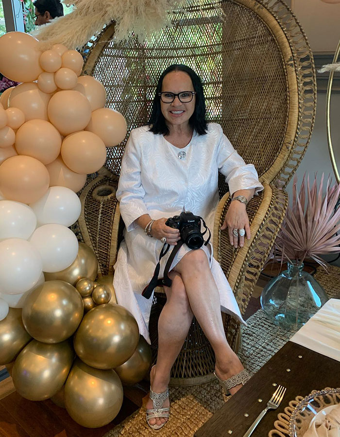 Woman smiling in a white dress holding a camera, sitting next to beige and gold balloons highlighting cancer diagnosis symptoms.