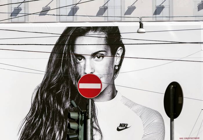Street photo showing a funny coincidence with a no entry sign perfectly covering a woman's mouth in a large Nike mural.