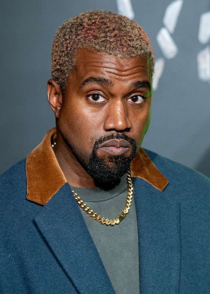 Kanye West in a blue coat and gold chain, showing a serious expression as fans react to public betrayal list involving daughter North.