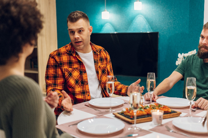Man in orange plaid shirt joking at dinner table with twin brother and woman about trading wife for younger woman.