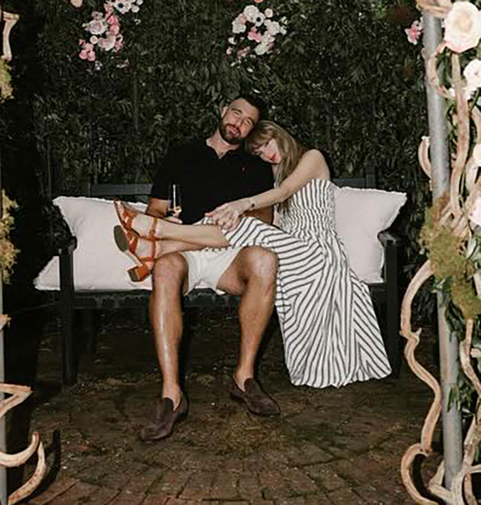 Travis Kelce and Taylor Swift sitting closely on a bench surrounded by flowers, capturing a tender moment together.