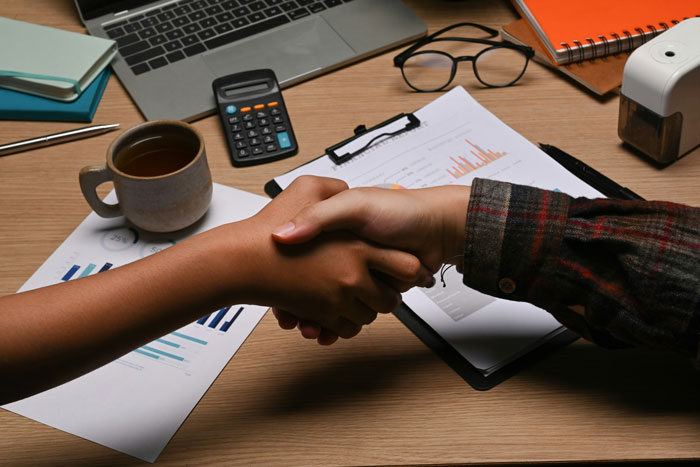 Two people shaking hands over a desk with charts and a laptop, symbolizing real-life cheat codes that work.