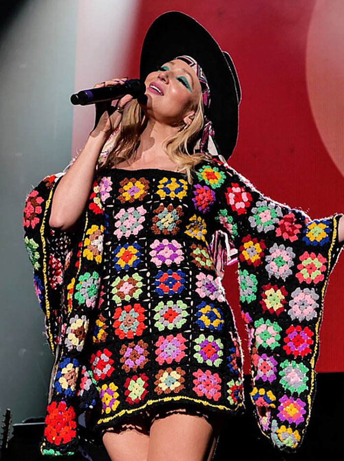 Female 90s star singing on stage wearing a vibrant crochet dress and wide-brimmed hat, showcasing confident style.
