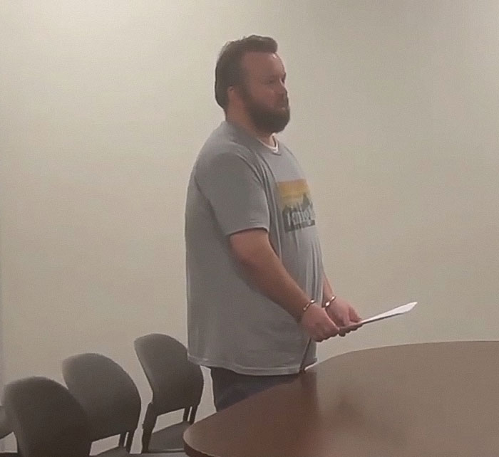 Man in handcuffs stands by a table in a plain room during a teacher arrest related to poop spray incident at school.