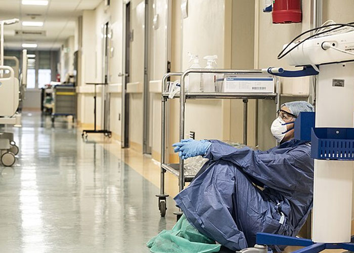 Healthcare worker in protective gear sitting exhausted in hospital corridor, highlighting societal damage from historical events.