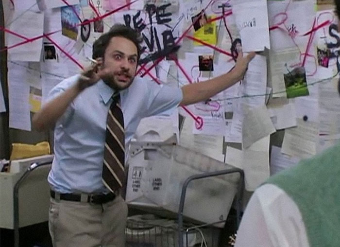 Man in a shirt and tie gesturing frantically in front of a chaos-filled office wall with papers and red string connections.