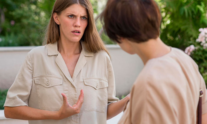 Two women having a tense conversation outdoors about a neighbor drilling holes in fence to spy.