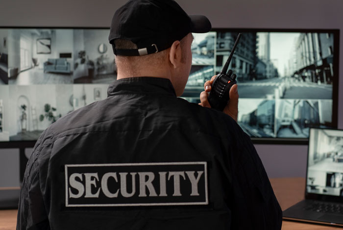 Security guard monitoring multiple surveillance screens and communicating through a walkie-talkie during a night shift.