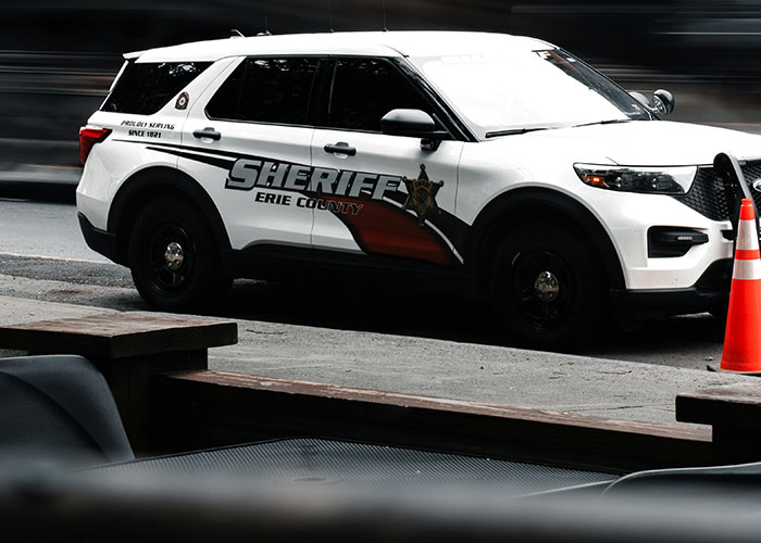 Sheriff patrol vehicle from Erie County parked on the street near an orange traffic cone during daytime, criminal fails theme.