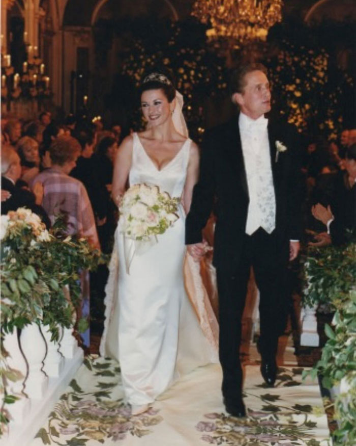 Catherine Zeta-Jones in a white wedding dress holding a bouquet, walking down the aisle with her groom in a formal setting.