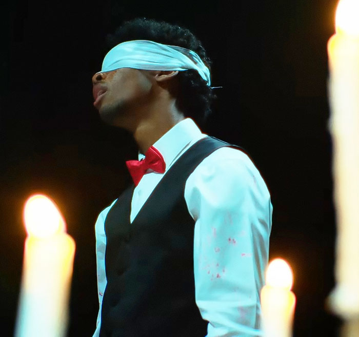 Blindfolded man dressed formally in vest and red bow tie, surrounded by lit candles in a dark setting.