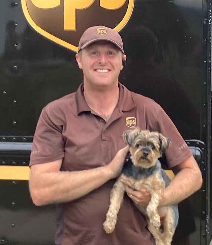 UPS driver in uniform smiling while holding a small dog in front of a UPS delivery truck logo