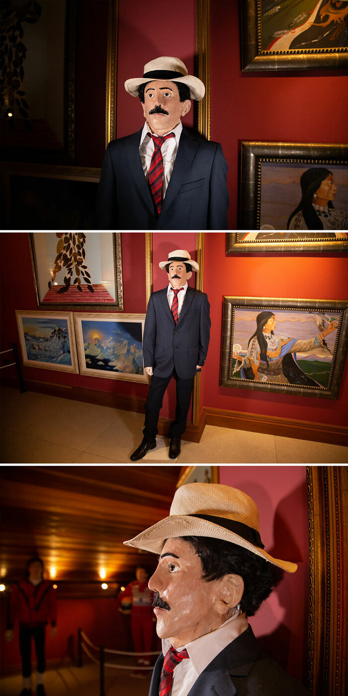 Wax sculpture of a famous figure dressed in a suit and hat, displayed in an art gallery with framed paintings.