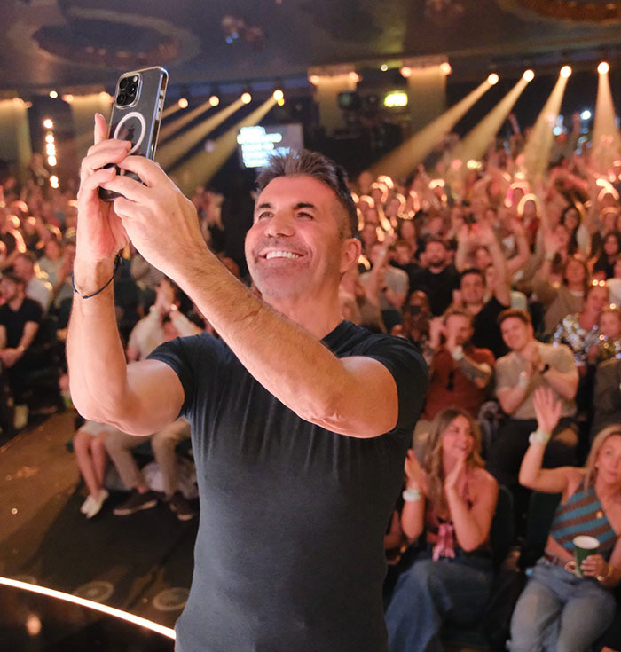 Simon Cowell smiling and taking a selfie with a large cheering crowd behind him after a bike accident survival.