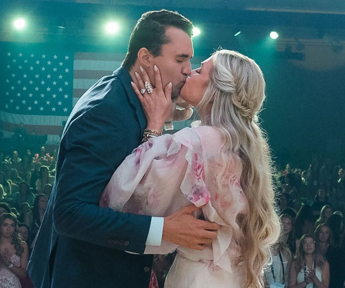 Elon Musk at a public event kissing a woman, with an American flag and audience in the background.