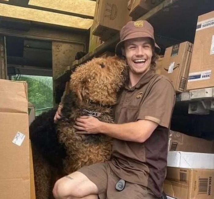 UPS driver in uniform happily hugging an adorable dog inside a delivery truck surrounded by packages and boxes.