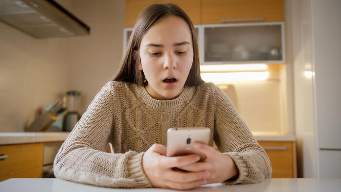 Young woman in a beige sweater sitting in kitchen, looking shocked at phone, reflecting the moment they realized they fell out of love.