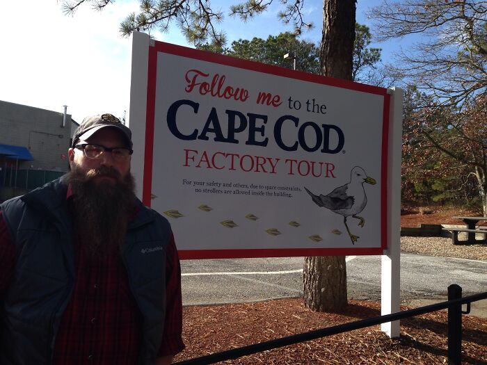 Man with a long beard standing next to a Cape Cod factory tour sign in a sunny outdoor setting, showcasing loophole use.