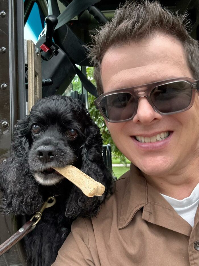 UPS driver smiling next to a black dog holding a bone, showcasing one of the adorable pets UPS drivers met on their route.