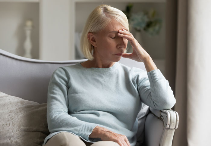 Middle-aged woman in a sweater sitting on a chair, looking distressed and holding her forehead in a living room.