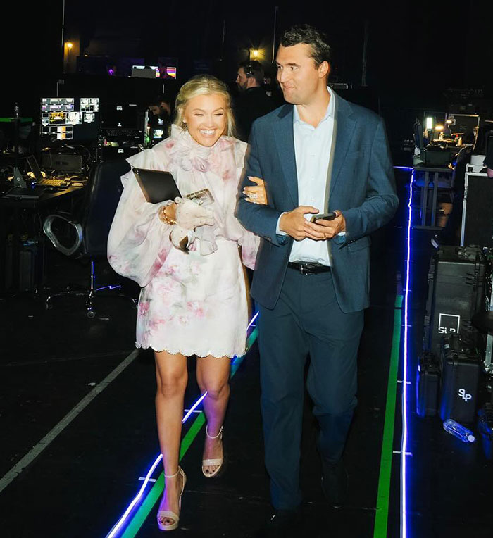 Charlie Kirk walking backstage with a woman, holding a phone and tablet in a dimly lit event setting.