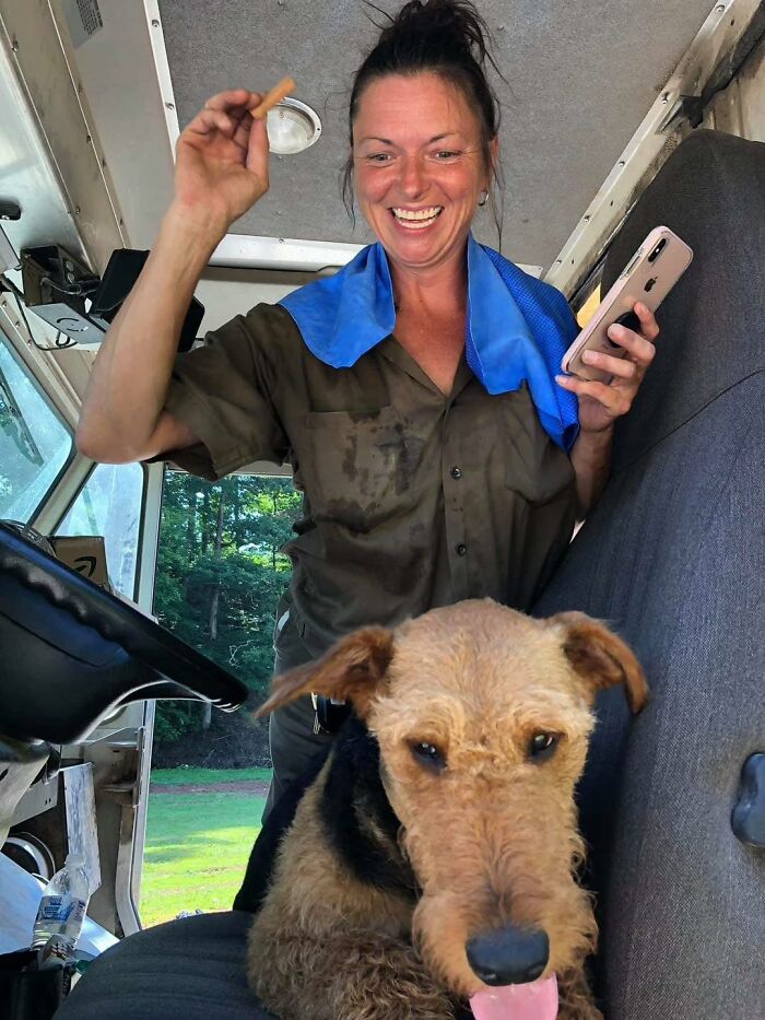 UPS driver with a phone smiling inside delivery truck with an adorable dog sitting on the seat nearby.