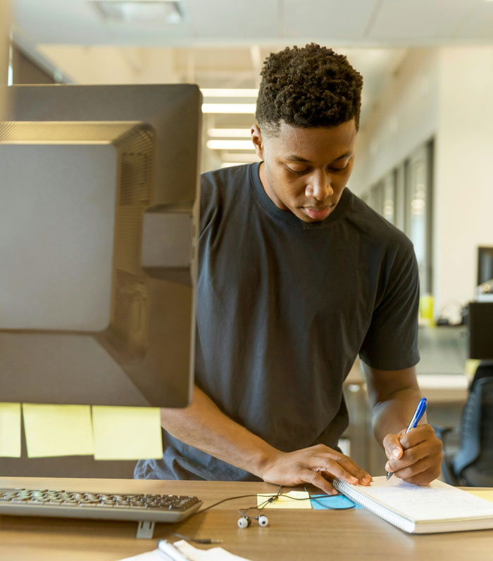 Young man writing notes at desk beside computer, illustrating productivity tips and real-life cheat codes for sleep benefits.