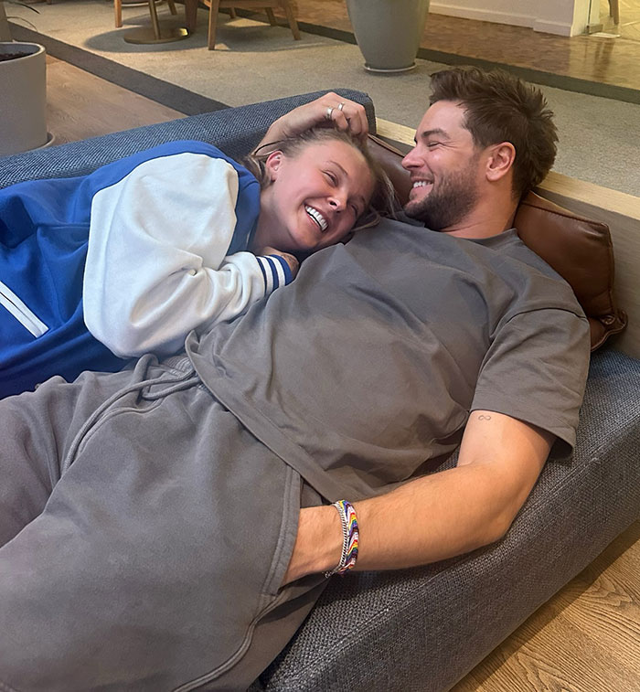 JoJo Siwa smiling and relaxing on a couch with a man, sharing a joyful and intimate moment together.