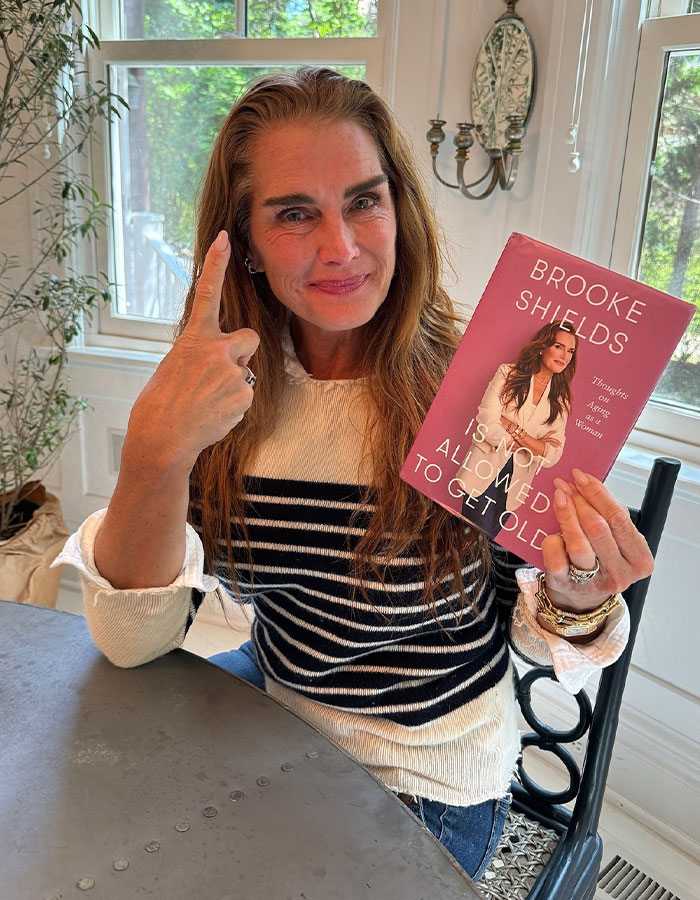 Brooke Shields holding a memoir, smiling and gesturing, representing raw and unfiltered confessions celebs shared in memoirs.
