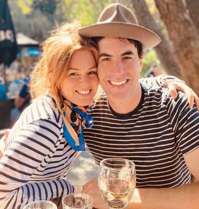 Sacha Baron Cohen smiling with a woman outdoors, both wearing striped shirts, enjoying a sunny day with drinks on the table. Sacha Baron Cohen smiling with a woman outdoors, both wearing striped shirts, enjoying a sunny day with drinks on the table.