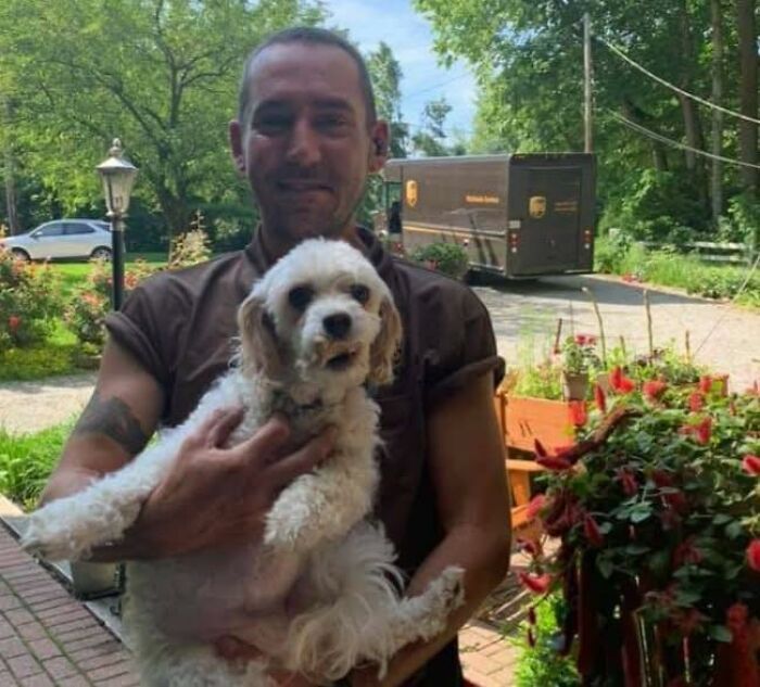 UPS driver holding an adorable small white dog outside a house with a UPS truck visible in the background on a sunny day.