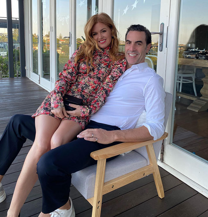 Sacha Baron Cohen smiling seated with woman in floral dress on outdoor patio with glass doors behind them. Sacha Baron Cohen smiling seated with woman in floral dress on outdoor patio with glass doors behind them.