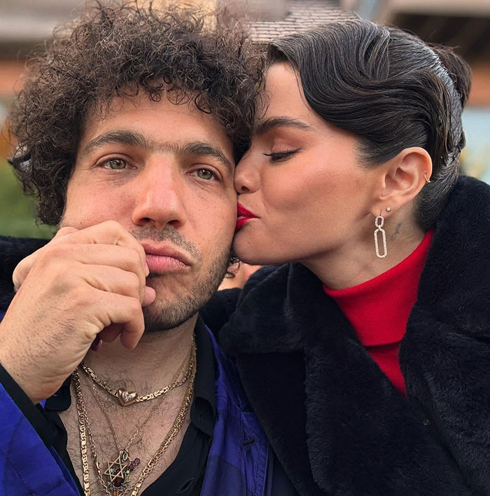 A woman in elegant earrings and red top kisses a man with curly hair wearing layered necklaces, close-up portrait.