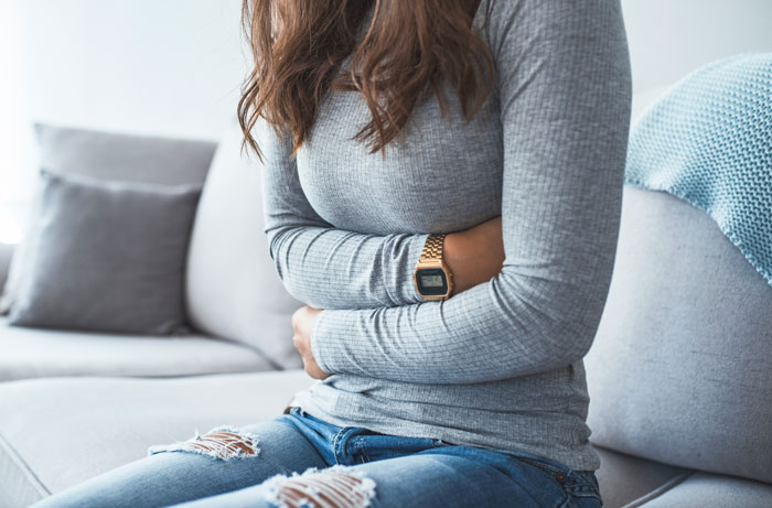 Young woman sitting on couch holding her stomach in distress, reflecting pain from betrayal and affair discovery.