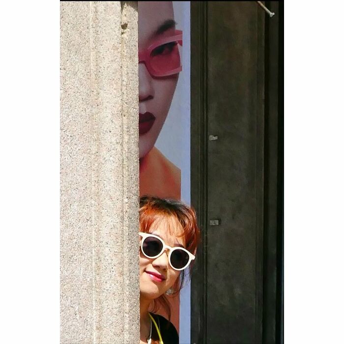 Woman wearing white sunglasses peeking from behind a wall, blending with a poster's face in a funny street photo coincidence.